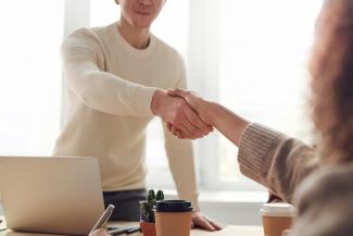 Man and woman shaking hands over a desk. On the desk is a laptop and a coffee cup.